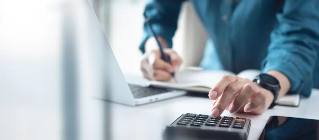 Person in a teal shirt uses a calculator on a desk while a laptop and notebook are nearby.
