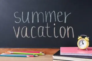 Chalkboard reading 'summer vacation' with a yellow alarm clock and school supplies on a desk.