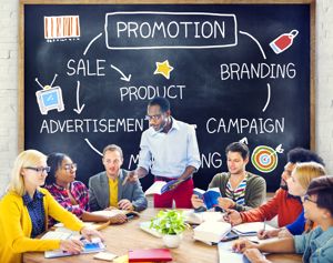 A diverse team stands in front of a blackboard covered with branding and advertising concepts. They are engaged in a lively conversation during a business conference, discussing strategies for a new advertising campaign. The setting reflects a collaborative effort in commerce and communication