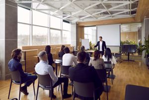 Team of people having class with business trainer. Group of male and female employees sitting at desks in modern office and listening to lecture by experienced teacher sharing knowledge and guidance