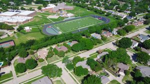 Aerial view of a middle school football field surrounded by residential houses, lush green trees, a swimming pool, and well-trimmed grassy front yards in an upscale suburban neighborhood in Dallas, USA.