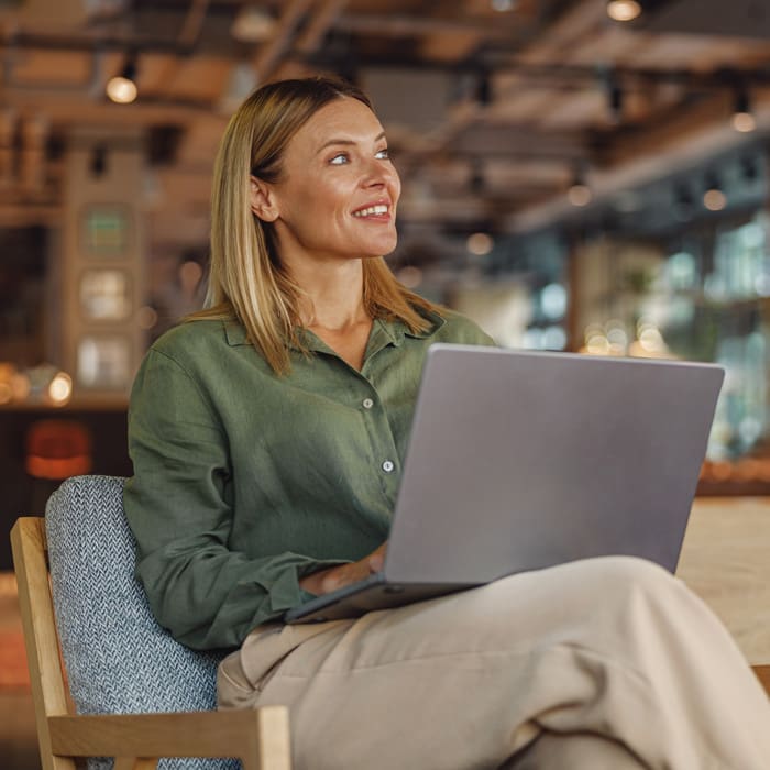 Woman in a green blouse sitting in a lounge area with a laptop on her lap, smiling and looking to the side.