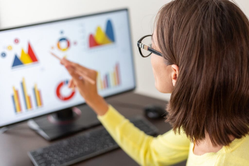 Woman wearing glasses points at colorful charts on a computer monitor during a data presentation.
