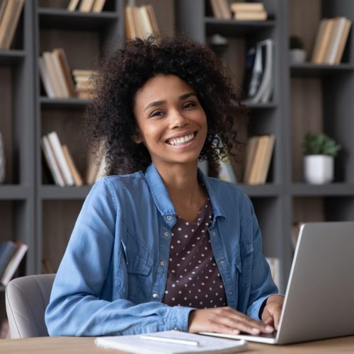 Happy millennial Black student girl in casual sitting at desk with laptop in library, looking at camera with toothy smile, doing research study. Head shot portrait